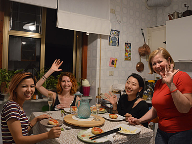 Internationale Sprachschueler sitzen gemeinsam mit ihrer Gastfamilie am Esstisch in der Küche, lachen und winken in die Kamera und erleben den Alltag während einer Sprachreise in Italien.