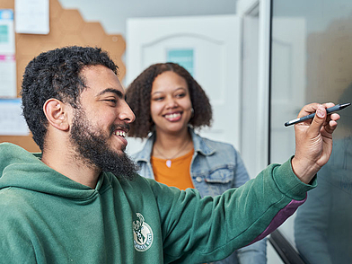 Ein Student schreibt während des Unterrichts an die Tafel, während eine andere Teilnehmerin in einer entspannten und motivierenden Atmosphäre aufmerksam zuhört.